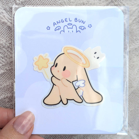 Sticker of a cute angel-like character with a halo, reaching for a star, on a light blue backing card, inside of a polybag, held by a hand.