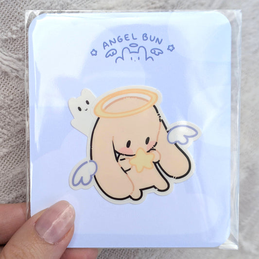 Sticker of an angel bunny held by a hand on a light blue card inside of a polybag with 'Angel Bun' text.