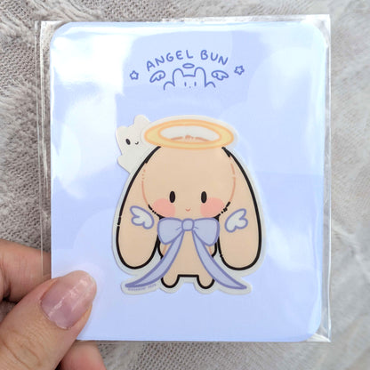 Sticker of angel bun character, with a light blue card backing, and polybag, held up by hand. Displays 'Angel Bun' text.