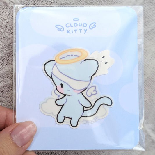 2 inch sticker of a cute cat-like character with a halo, on a light blue backing card, inside a polybag, held by a hand.